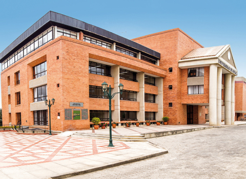 Edificio institucional de ladrillo con columnas, ventanales y plaza exterior en un campus educativo